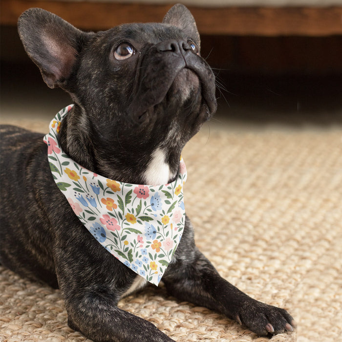 French Bulldog wearing a white floral bandana with pink, blue, and yellow flowers, resting on a woven rug.