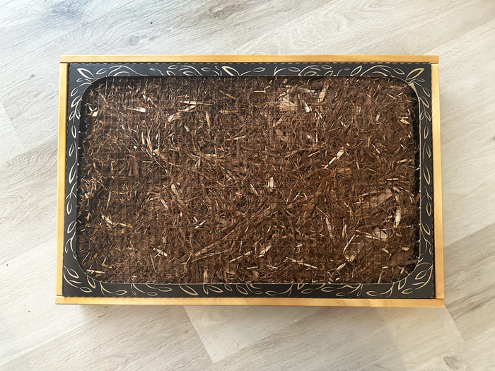 Top-down view of a Bark Potty disposable pad seated inside a wooden tray sleeve on a wood floor.