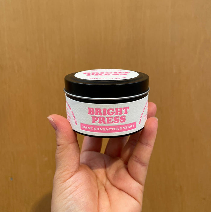 Hand holding a candle tin labeled 'Bright Press - Mane Character Energy' against a wooden background